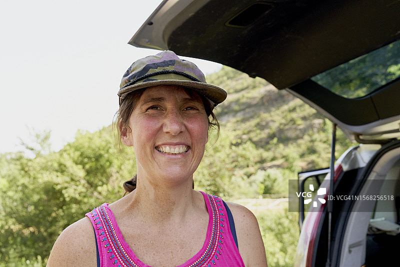 戴帽子、穿休闲服装的女人站在打开的汽车后备箱旁，面带微笑或笑声地看着镜头，正面视角图片素材
