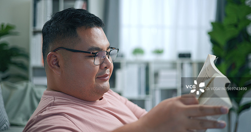 快乐的年轻超重男子在客厅沙发上看书休息图片素材