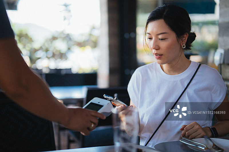 年轻亚洲女性在餐厅用智能手机进行移动支付，在刷卡机上扫描并支付账单，进行快速便捷的非接触式支付。NFC技术，轻触即付概念图片素材