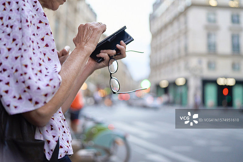 在人行道上使用手机并拿着眼镜，等待出租车回家的老年妇女图片素材