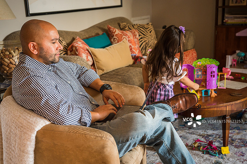 父亲在家用笔记本电脑工作，女儿在家里玩耍图片素材