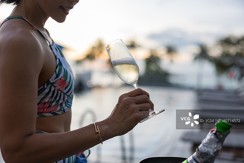 酒店泳池的夏日氛围：微笑的女人在日落时分举着酒杯，热带假期图片素材