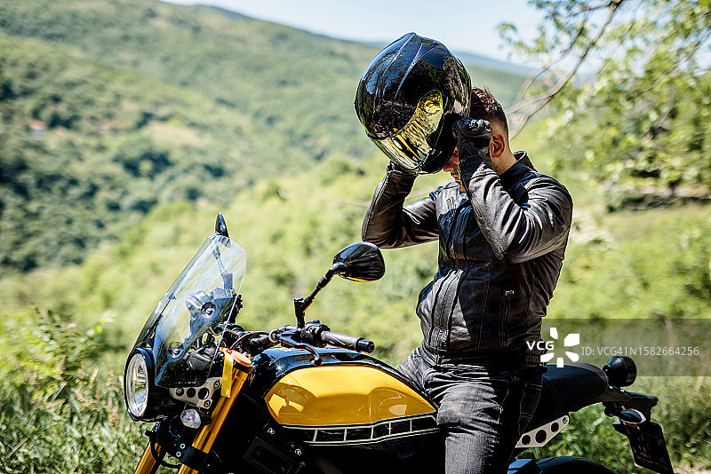 在乡村公路骑行休息后戴上安全头盔的英俊摩托车手图片素材