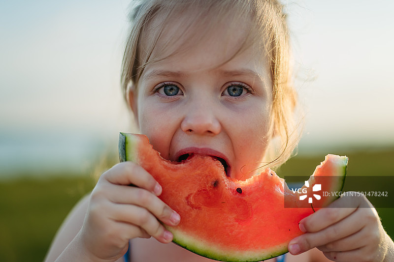 在海滩上吃西瓜的可爱小女孩肖像。在假日和暑假期间吃甜美的水果。图片素材