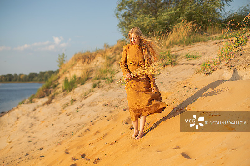 穿着黄色连衣裙，在河岸边休息的金发女人。日落光。夏天。长发。沙子。心碎图片素材