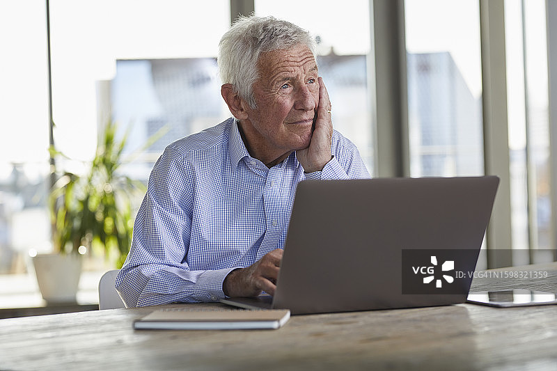 沉思的老年男人肖像，坐在桌旁用笔记本电脑看着远方图片素材