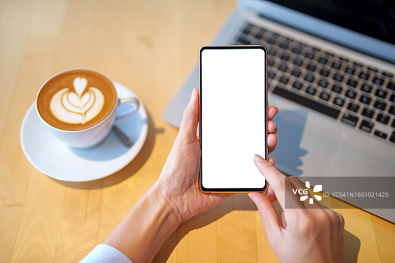 女性手持空白屏幕智能手机样机，背景为笔记本电脑和咖啡杯图片素材