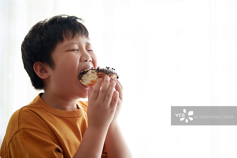 一名亚洲男孩站在窗边咬了一口巧克力甜甜圈，旁边有额外的空白内容图片素材