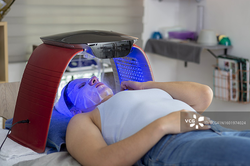 在美容师办公室接受LED光疗的戴着面膜的年轻墨西哥女人图片素材
