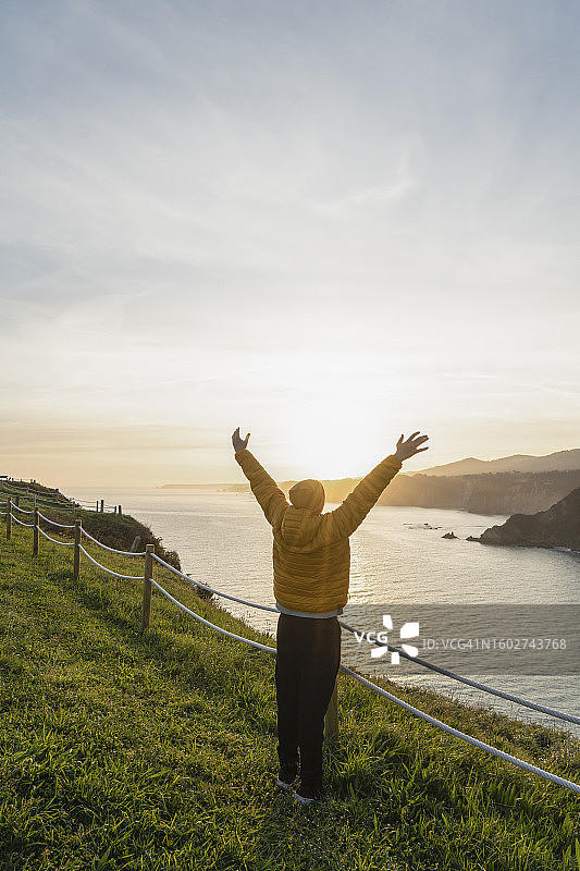 穿着雨衣戴着黄色帽子在海滩悬崖边张开双臂的青少年图片素材
