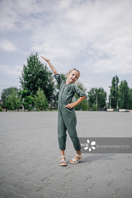 身穿绿色连体裤和凉鞋的苗条女孩站在铺砌的院子里，举起手图片素材