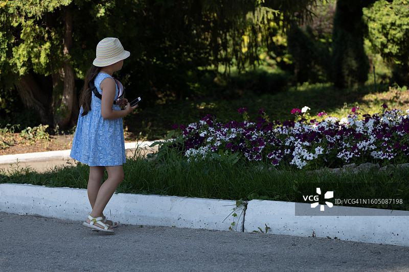 可爱的小女孩拿着手机走过花坛，迷人的花朵图片素材