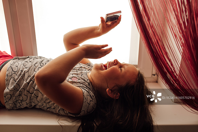 快乐的小女孩仰卧在窗台上，举着智能手机在脸上方，无法控制地笑着图片素材