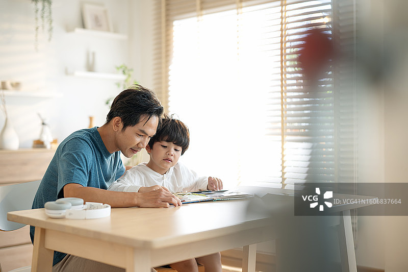 父母在家辅导孩子手写作业，提升技能和情商。侧面拍摄儿子在客厅里在父亲的指导下用平板电脑完成作业。图片素材