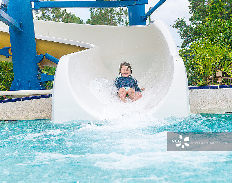 在水上乐园或度假酒店，女孩从水滑梯滑入泳池图片素材