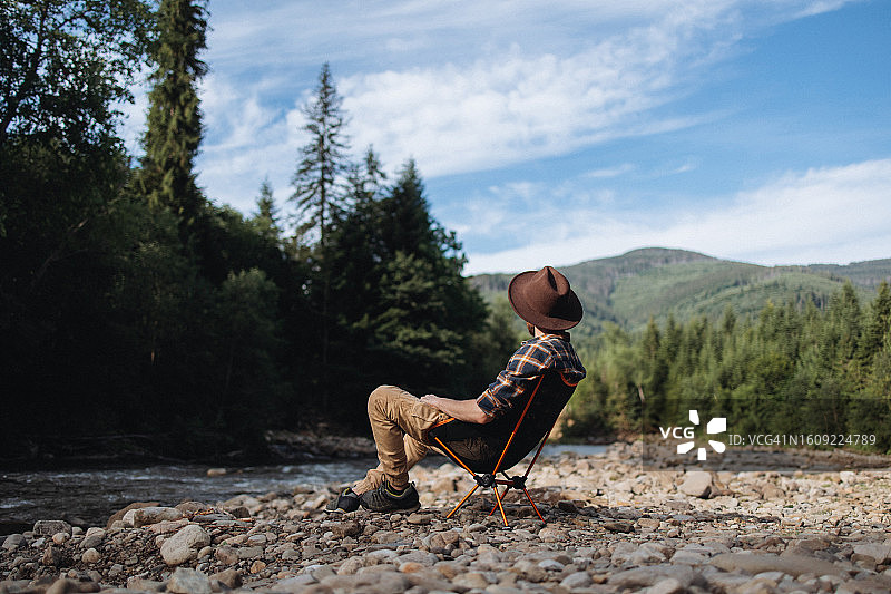 戴帽子的孤独男人坐在山间河流附近的椅子上休息图片素材