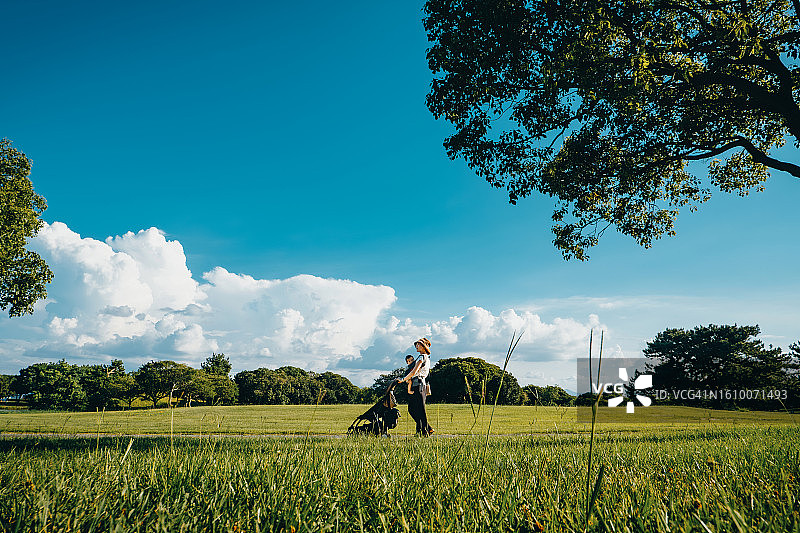 年轻的亚洲母亲用婴儿背带背着她的女儿在阳光明媚的自然公园的草地上散步，家人享受大自然，母女时光，爱和关怀，家庭、儿童和育儿理念图片素材
