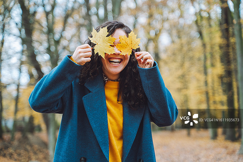 笑容满面的女人拿着枫叶遮住脸庞：一位有趣的黑发女人在秋季公园或林地里，用一片大枫叶俏皮地遮住脸庞，玩得很开心。图片素材
