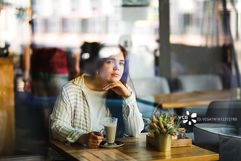 咖啡馆里的孤独：悲伤美丽的超重长发女人在窗边寻求慰藉，看着镜头图片素材
