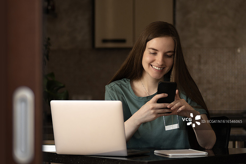 在家使用笔记本电脑和手机工作的长发女人，背景是厨房和客厅图片素材
