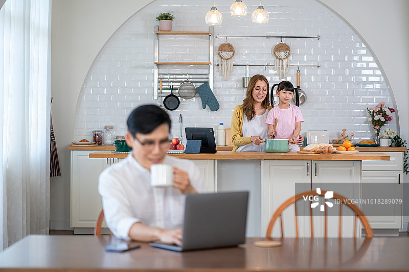 男人在家中厨房的桌旁使用笔记本电脑工作，家人在旁图片素材