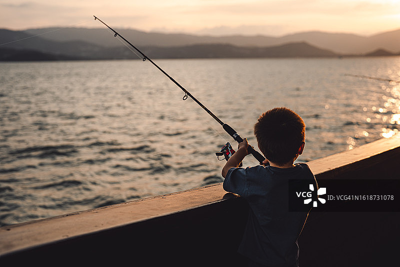 可爱小男孩在船上钓鱼，手持钓竿，沐浴在金色夕阳下。特写，后视图。图片素材
