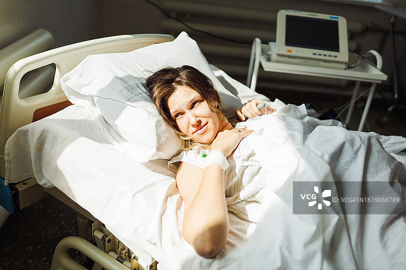 迷人的孕妇穿着医院病号服，躺在阳光明媚的医院病床上图片素材