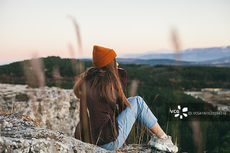 年轻女子戴着无檐小便帽坐在秋季悬崖顶上，欣赏日落时分美丽的山景图片素材