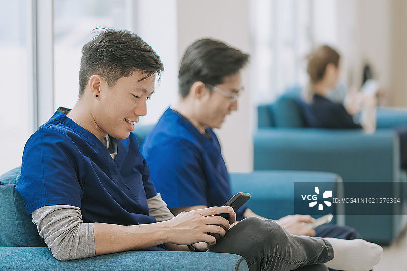 快乐的亚洲华人男护士穿着医疗服坐在沙发上阅读智能手机短信图片素材