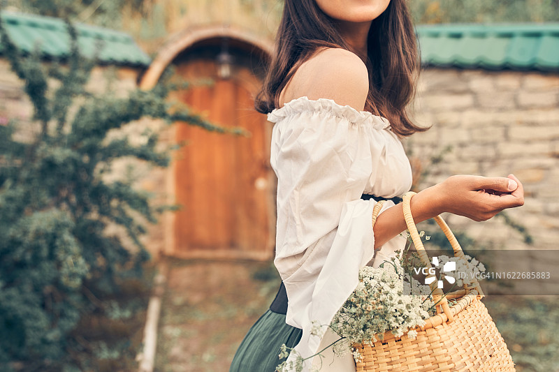 一个童话般的，无法辨认的女人站在森林里她家附近，一个穿着戏服的女人，手里拿着一篮子鲜花，正在眺望远方。为你的故事准备的中世纪服装。带有复制空间的图书封面设计。图片素材