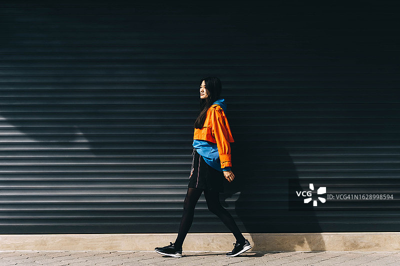 穿着色彩鲜艳服装的年轻亚洲女性走在黑色墙壁旁，生活方式和青年文化图片素材