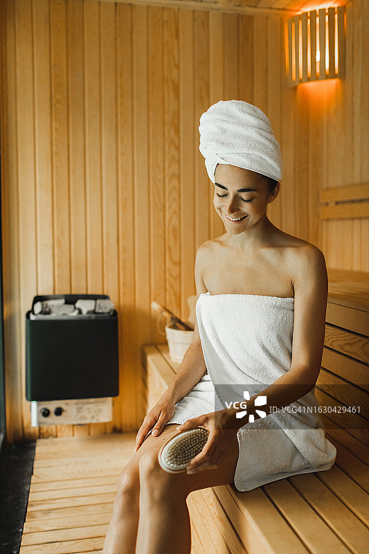 在桑拿房休息，用天然木刷按摩腿部的快乐年轻女人图片素材