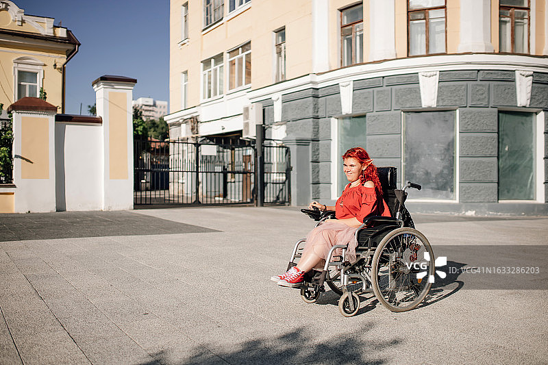 快乐的年轻红发女士坐在轮椅上在阳光明媚的城市街道上行驶图片素材