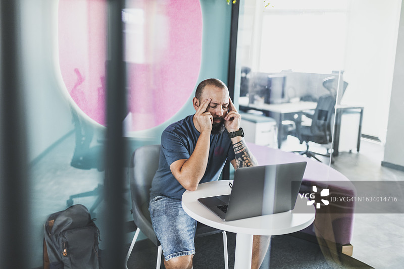 疲惫的程序员坐在共享办公桌旁，使用笔记本电脑图片素材