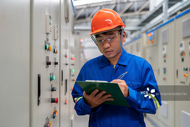 电气工程师在开关设备室内检查控制面板上的电气系统，并在剪贴板上记录。在受控环境中维护电气系统完整性的仔细检查。图片素材