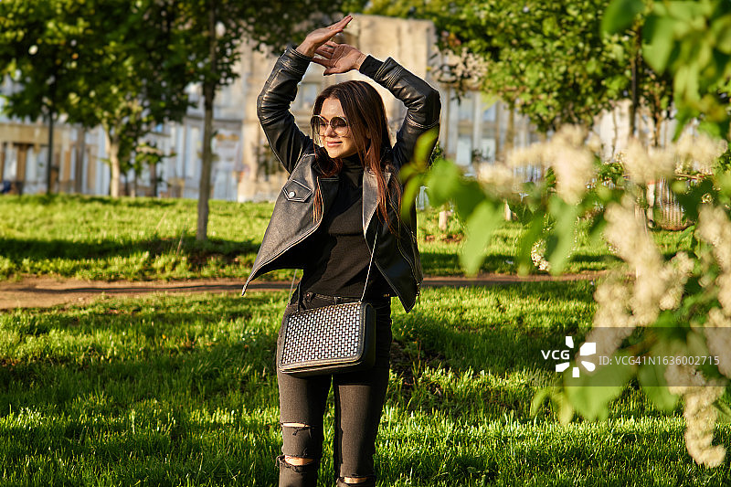 有魅力的年轻千禧一代女性在街上。她站在美丽的绿色盛开的树木中，享受生活和阳光。图片素材