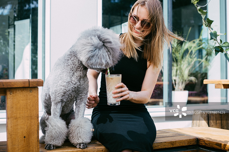 快乐的爱狗女士坐在路边咖啡馆的长椅上，与蓬松的灰色贵宾犬一起喝奶昔图片素材