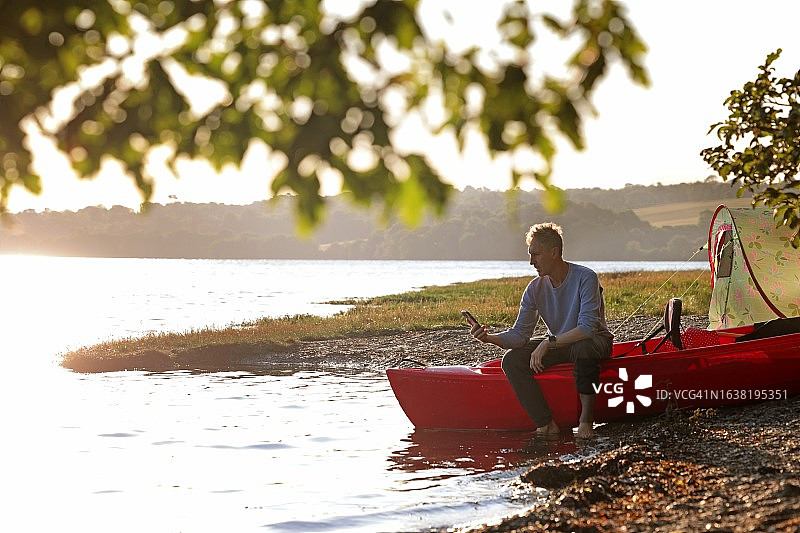 男人坐在河边的独木舟上看手机图片素材