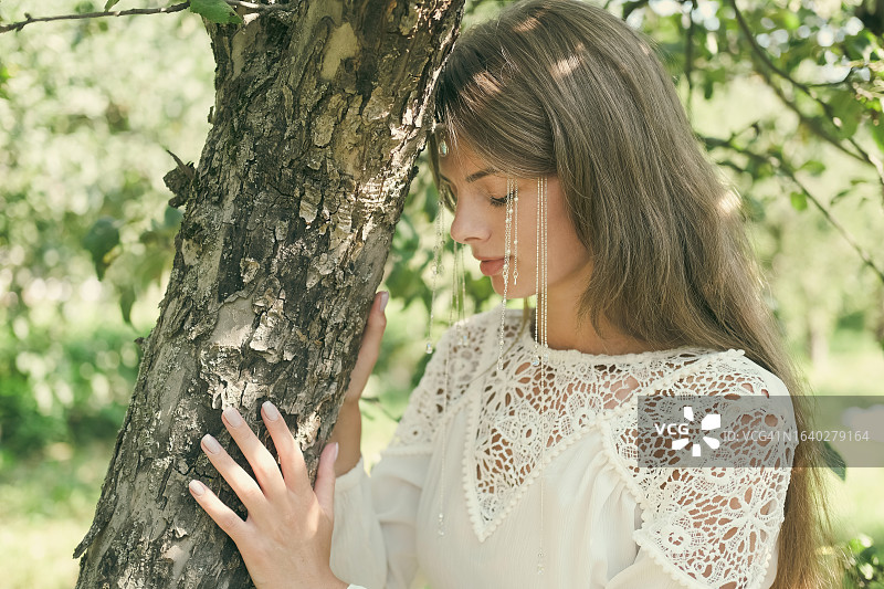 身穿白色连衣裙的年轻女人在户外触摸树木。图片素材