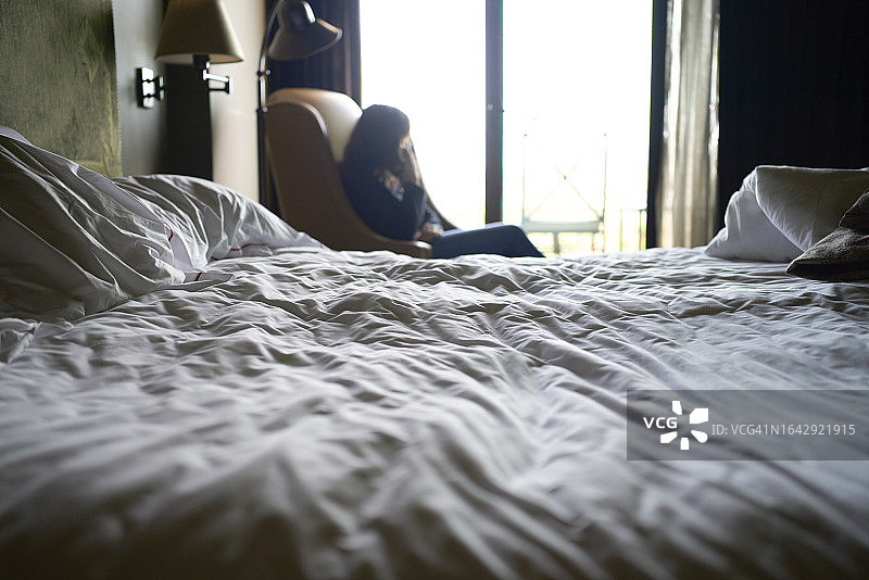 一张不整洁的床，背景中一位女士坐在家具上，两盏灯和一个被阳光照亮的玻璃门图片素材