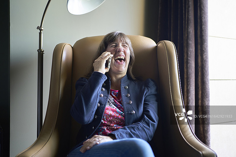 穿着蓝色西装，坐在米色沙发上，大声笑着打电话的女人图片素材