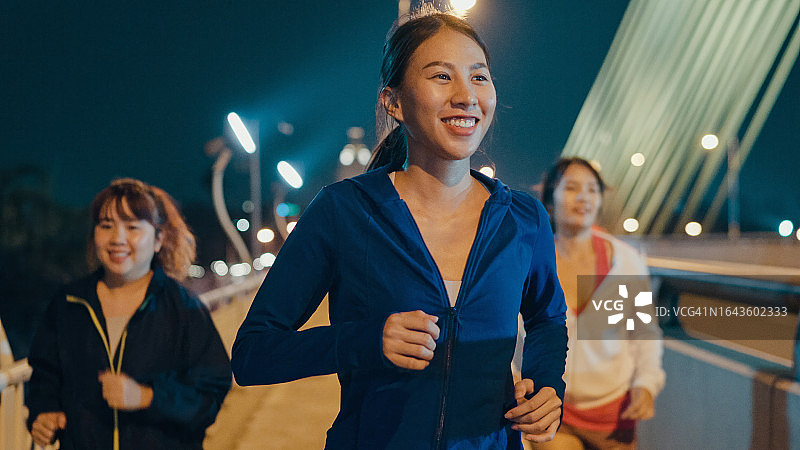 夜间城市街道桥上，多种族年轻运动型亚洲女性穿着运动服跑步，享受有氧运动。跑步俱乐部和户外运动概念。图片素材
