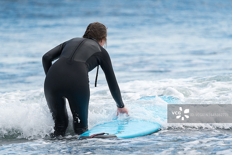 穿着潜水服、手持冲浪板的年轻女运动员走向太平洋海浪的背影图片素材