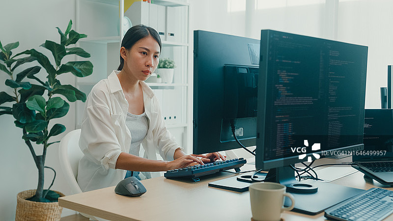 年轻的亚洲女性软件开发人员使用电脑编写代码，坐在办公桌前，多个屏幕在办公室工作。程序员开发。图片素材