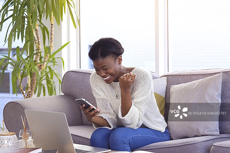 交易成功：快乐的黑人女性在沙发上用手机查看电子邮件或在线获胜。快乐的远程工作者在手机上阅读奖金或成就信息图片素材