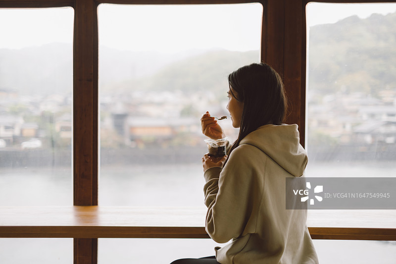 年轻亚洲女性在日本咖啡馆品尝抹茶冰淇淋，透过咖啡馆的窗户欣赏宇治风景的背影。美食、放松、旅行、休闲和度假概念图片素材