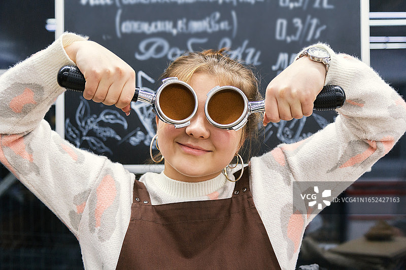 在咖啡店用咖啡手柄遮住眼睛的有趣年轻咖啡师女孩图片素材