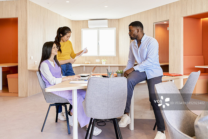 三位多种族同事坐在办公桌旁交谈并微笑的侧视图图片素材