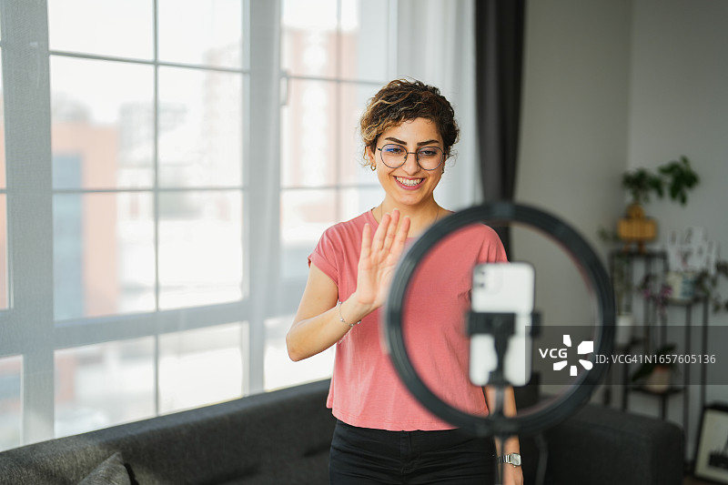 坐在客厅沙发上录制社交媒体视频，向镜头挥手的年轻女人图片素材