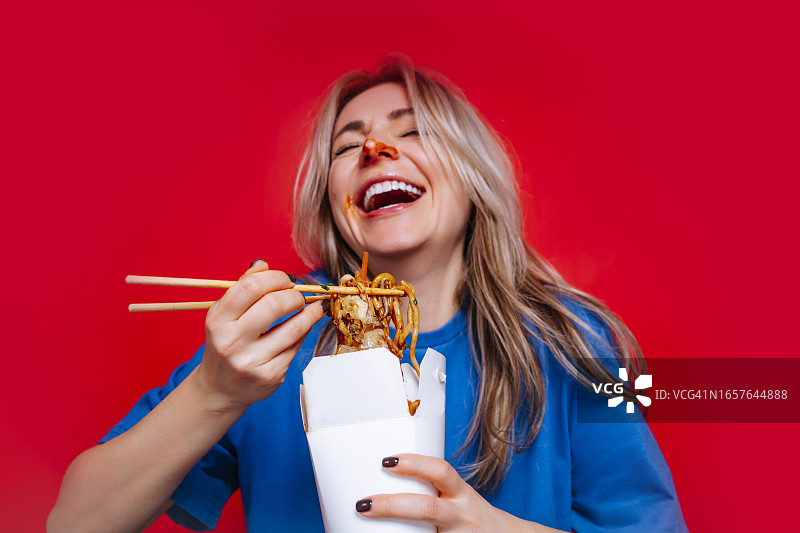 快餐食品：盒装即食面条。一位金发女士用筷子吃着美味的辣味面条，鼻子沾满了酱汁，开怀大笑图片素材